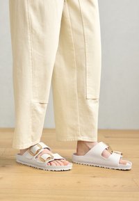 Sandali bianchi a due cinturini realizzati in materiale morbido, con fibbie dorate e una suola testurizzata. Indossati con pantaloni larghi color crema.