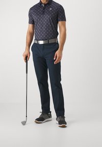 Marinblå polo skjorta med geometriskt mönster, kombinerad med mörka byxor. Svarta och grå idrottsskor. Håller en golfklubba med ett silverfärgat bälte.