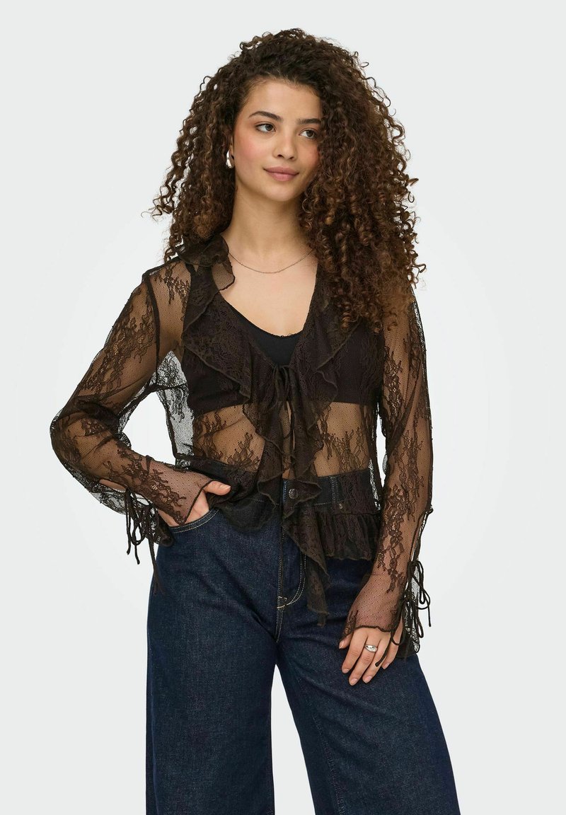 Femme aux cheveux bouclés portant un chemisier en dentelle noire transparente sur un haut noir et un jean taille haute bleu foncé, debout avec une main dans la poche.