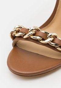 Sandal in pelle marrone con una larga fascia decorata da una catena dorata. Texture liscia, punta rotonda e suola piatta.