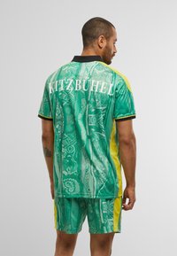 Mann mit kurzen Haaren, der ein grün gemustertes Sportoutfit mit gelben Seitenpanelen trägt und "KITZBÜHEL" auf dem Rücken aufgedruckt hat.