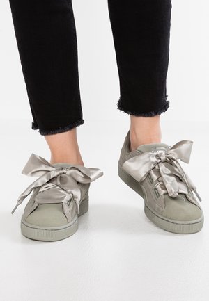 Paar hellgraue Sneakers mit breiten Satinband-Schnürsenkeln, getragen zu schwarzen Hosen mit ausgefranstem Saum auf weißem Boden.