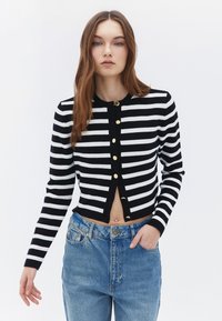 Randig svart och vit cardigan med rund halsringning, guldknappar och en kort design. Kombineras med högmidjade blå denimjeans.