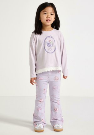 Nuori tyttö seisoo laventelivärisessä asussa, jossa on kakkuaiheisia kuvia, kuten kakkuaiheinen kuvitus paidassa ja erilaisia jälkiruokia housuissa.