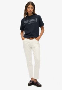 T-shirt en coton marine avec un graphisme "SUPERDRY ATHLETICS", associé à un jean blanc et une ceinture tressée marron. Le modèle porte des mocassins foncés.