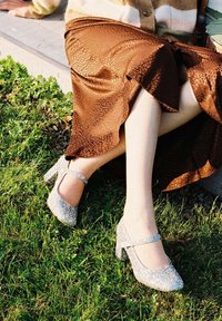 Zapatos de tacón alto plateados con brillo, con puntera redonda y correa al tobillo, combinados con una falda marrón texturizada, sobre césped verde.