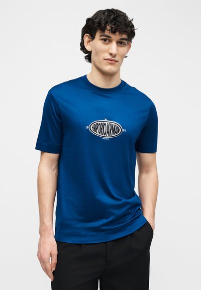 Νεαρός άνδρας με σκούρα σγουρά μαλλιά, ντυμένος με μπλε μπλουζάκι Emporio Armani και μαύρο παντελόνι, στέκεται με το ένα χέρι στην τσέπη, πάνω σε λευκό φόντο.