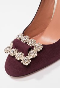 Chaussure à talon haut en daim bordeaux, ornée d'une grande broche embellie de strass sur l'avant, avec une doublure intérieure lisse et un bout arrondi.