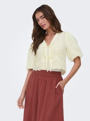 Femme aux longs cheveux bruns, portant un chemisier en dentelle jaune pâle à œillets avec manches bouffantes et un pantalon taille haute couleur rouille, main dans la poche.