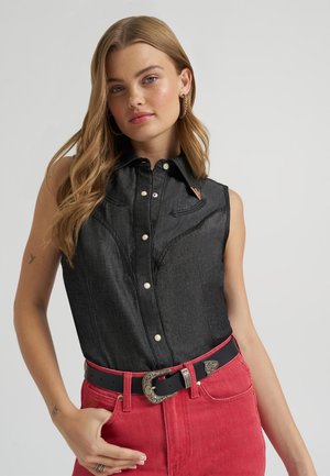 Nuori nainen, jolla on pitkät vaaleat hiukset ja joka on pukeutunut vyötäröään korostaviin punaisiin housuihin mustalla vyöllä ja mustaan hihattomaan farkkutoppiin, poseeraa yksiväristä taustaa vasten.