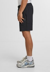 Svarte sportshorts med snøring i midjen, med sidelommer og en logo. Kombinert med grå og blå løpesko.