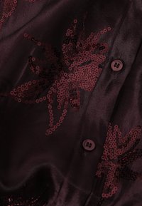 Blouse bourgogne avec une texture lisse, ornée de motifs floraux en sequins dans des teintes assorties et de deux boutons près du col.