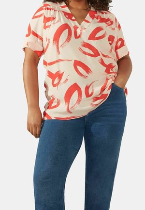 Frau trägt cremefarbene kurzärmelige Bluse mit rotem abstraktem Pinselstrichmuster und blaue Jeans, mit einer Hand in der Tasche.