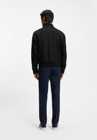 Giacca bomber nera con orlo e polsini a coste; tessuto liscio; vista posteriore con dettaglio del logo al colletto, abbinata a pantaloni blu e sneakers.