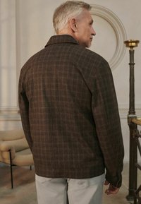 Brown plaid jacket with a collar, featuring textured fabric and a relaxed fit. Back view shows clean lines and simplicity in design.