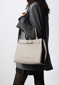 Sac à main en cuir beige avec une forme structurée, une poignée supérieure et des garnitures en or. Présente une fermeture plate et une surface texturée. Posé sur un fond gris.