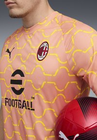 Rožinė Puma futbolo marškinėliai su geltonu geometriniu raštu, su apvaliu logotipu ir juodu tekstu. Laiko raudonai juodą futbolą.