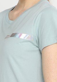 Detailní záběr osoby v světle modrém tričku s horizontálním lesklým stříbrným odrazným pruhem přes hruď.