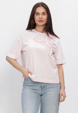 Vrouw met lang donker haar draagt een lichtroze top met korte mouwen en bloemenapplicatie, gecombineerd met lichtblauwe spijkerbroek, staand tegen een effen witte achtergrond.