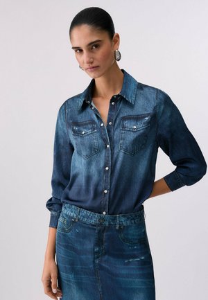 Mujer con el pelo peinado hacia atrás, llevando pendientes plateados, una camisa vaquera azul oscuro abotonada y una falda vaquera a juego sobre un fondo liso.