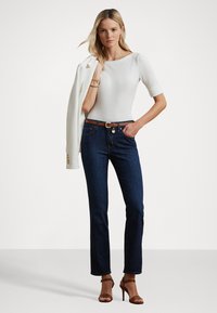 Blauwe jeans met een slim fit, gecombineerd met een witte aansluitende top en een crèmekleurige blazer. Bruine riem met hardware-detail en bruine haksandalen.