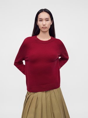 Femme aux longs cheveux foncés portant un pull rouge et une jupe plissée beige debout devant un fond clair uni.