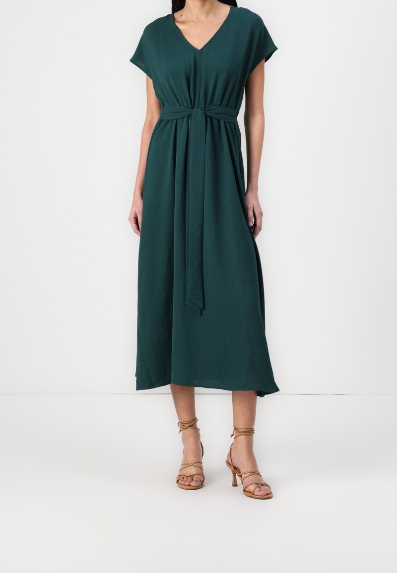 Femme portant une robe midi vert foncé à col en V avec ceinture à la taille et des sandales beiges à talons hauts à fines brides, debout sur un fond blanc.