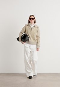 Suéter de malha bege com colarinho com fecho, combinado com uma camisa branca e calças largas brancas. A segurar uma carteira preta, usando óculos de sol escuros.