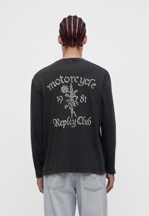 Person med flätat hår som bär en svart långärmad tröja med en kniv- och rosdesign och texten "motorcycle 1981 Replay Club" på ryggen.