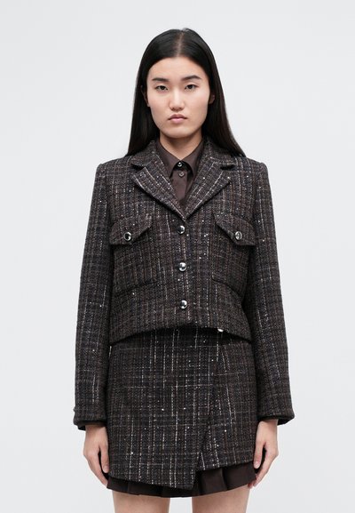 Pruun troopiline jakk hõbedaste detailidega, lühendatud lõikega, kahekordsete rinnataskutega ja nööpidega kinnitatav, kombineeritud tumeda särgi ja sobiva seelikuga.