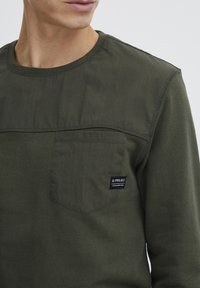 Olive green long-sleeve shirt made of cotton, featuring a textured chest panel and a small label on the pocket.