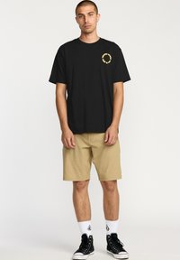 T-shirt en coton noir à manches courtes avec un logo jaune sur la poitrine gauche, associé à un short kaki et des baskets montantes noires.