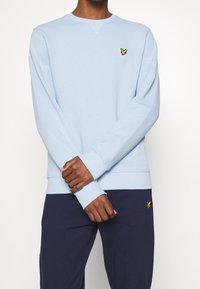 Sudadera azul claro de tejido suave, con mangas largas y escote redondo. Incluye un pequeño logo amarillo bordado en el pecho.