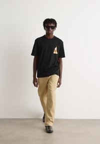 Svart t-skjorte med korte ermer som har et lite, fargerikt grafisk motiv på brystet, kombinert med beige bukser og svarte slip-on sko.