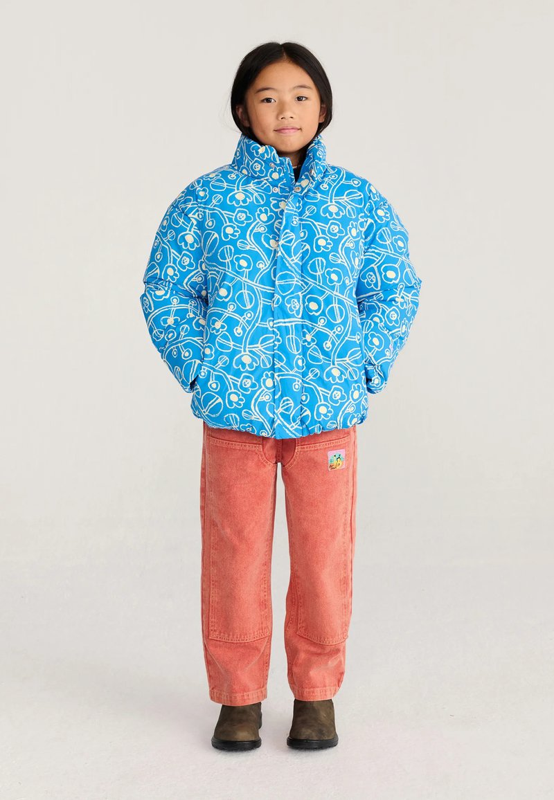 Enfant debout les mains dans les poches, portant une doudoune bleue à fleurs, un pantalon rouge avec un patch et des bottes foncées sur un fond uni.