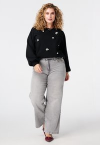 Blonde vrouw met krullend haar die een zwarte trui met zilveren bloemenapplicaties, grijze wijde jeans en bordeauxrode hakken draagt, terwijl ze vooruit loopt.