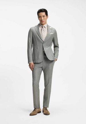 Uomo con abito grigio chiaro, camicia beige, cravatta a righe, fiore beige sulla giacca e scarpe eleganti marroni, in piedi con una mano in tasca.