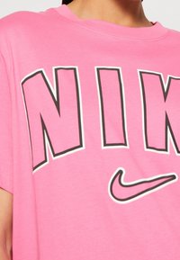 Rosa bomullströja med stora svarta bokstäver som stavar "NIKE" med vita konturer och en liten svart Nike-swooshlogotyp. Kort ärm.