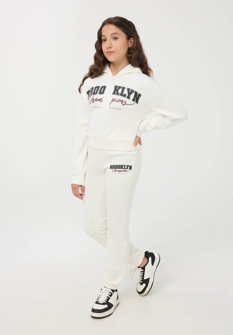 Biała bluza z kapturem na zamek z napisem "BROOKLYN", zestawiona z dopasowanymi dresami. Czarne akcenty i sneakersy dopełniają stylizację. Gładka tekstura materiału.