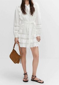 Robe blanche avec des accents à volants, des boutons et une ceinture en tissu. Associée à un sac à main en daim marron et des sandales noires à lanières.