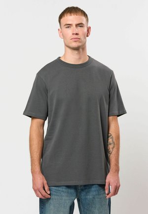 Joven con el pelo corto y castaño, vestido con una camiseta lisa de color gris oscuro y vaqueros azules, de pie frente a un fondo blanco.