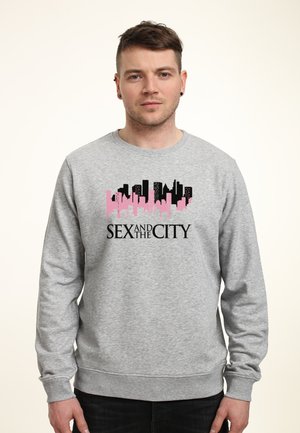 Man draagt een grijze sweater met een roze en zwarte stadsgezicht en de tekst "SEX AND THE CITY" aan de voorkant, staand tegen een effen achtergrond.