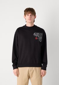 Sudadera negra de algodón, con cuello redondo y mangas largas. El bolsillo frontal presenta un patrón de paisley blanco y un logotipo rojo.