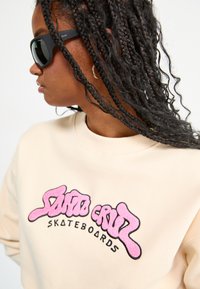 Felpa crema con ricamo nero e rosa bubblegum "Santa Cruz Skateboards". Il modello indossa occhiali da sole neri; capelli intrecciati visibili.