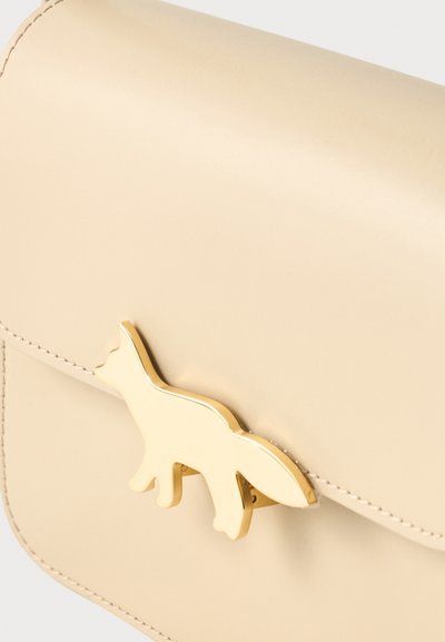 Τσάντα από δέρμα χρώματος κρέμα με μεταλλικό κούμπωμα σε σχήμα αλεπούς σε χρυσό χρώμα στην εμπρόσθια πτερύγιο, που δείχνει λεία υφή και λεπτομερή ραφή.