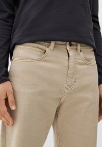 Jeans de algodón beige con corte recto, bolsillos frontales y cierre de cremallera. Tela texturada con detalles de costura visibles.
