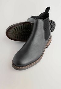 Fekete bőr Chelsea csizma elasztikus oldalsó panelek, texturált talpárok és barna varrással díszített középtalp részletekkel a világos háttér előtt.