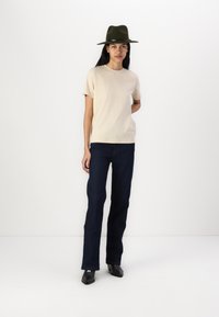 Beige kortermet t-skjorte, mørkeblå utsvungne jeans og svarte sko med firkantet tå. Grønn bredbremmet hatt som tilbehør.