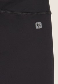Leggings negros con una textura suave, que presentan un pequeño parche de logo brillante. Las costuras son visibles a lo largo de la cinturilla y las costuras.