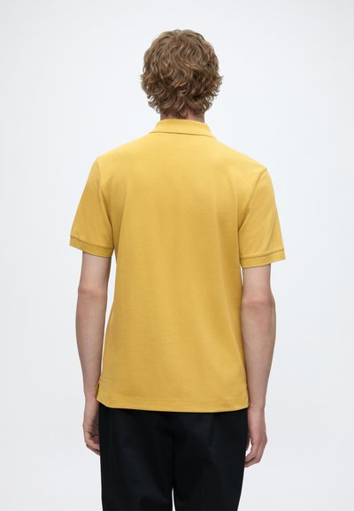 Jeune personne aux cheveux bouclés portant un polo jaune à manches courtes et un pantalon noir, debout de dos sur un fond uni.